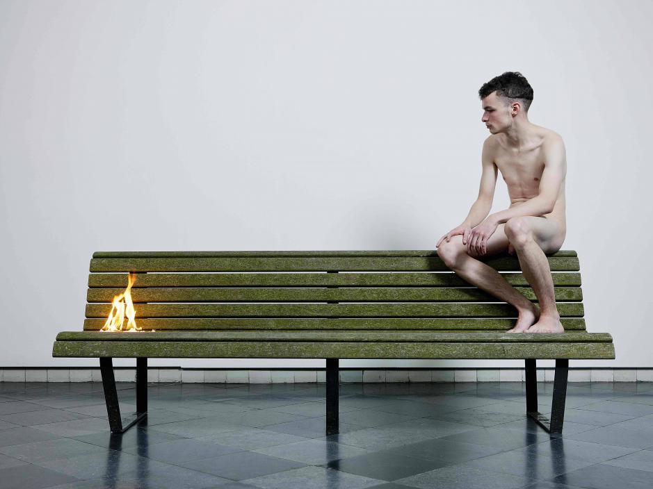 A nude man sitting on a bench while the other side is on fire