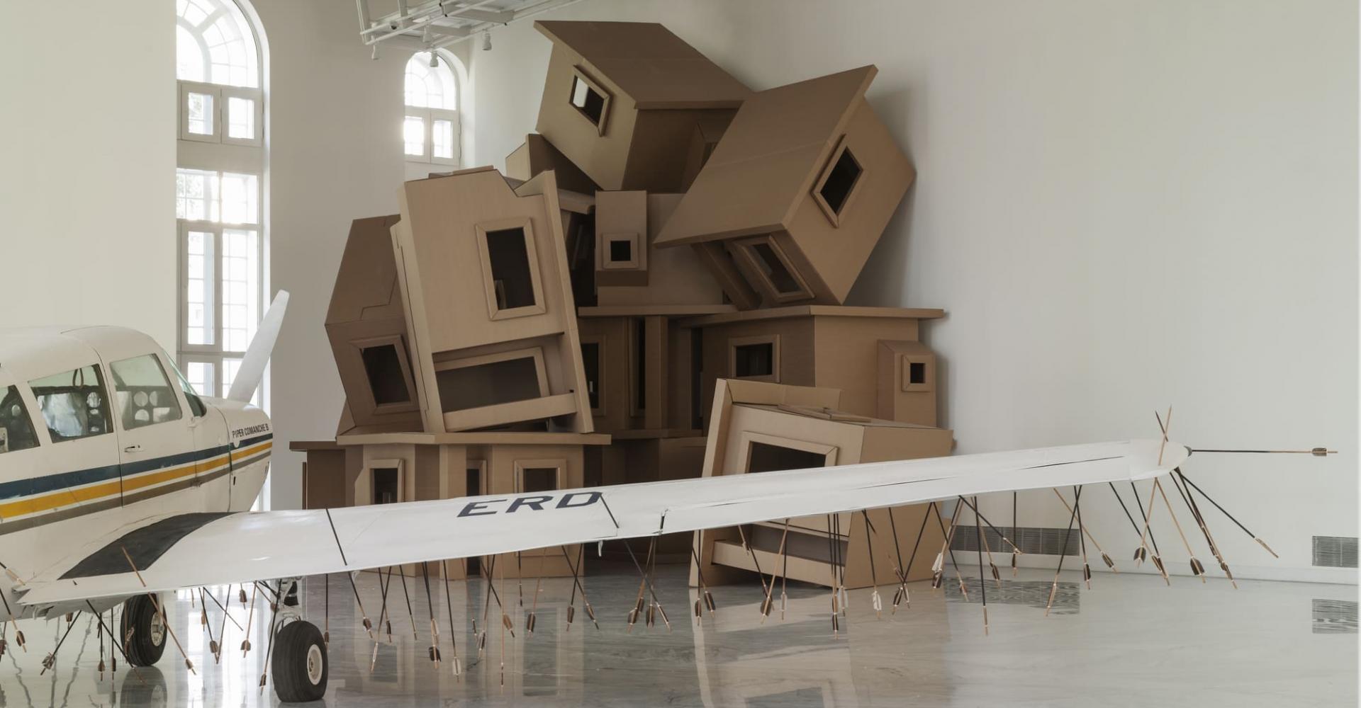 Cardboard houses stacked with a plane whose underbelly is covered in arrows in the foreground.